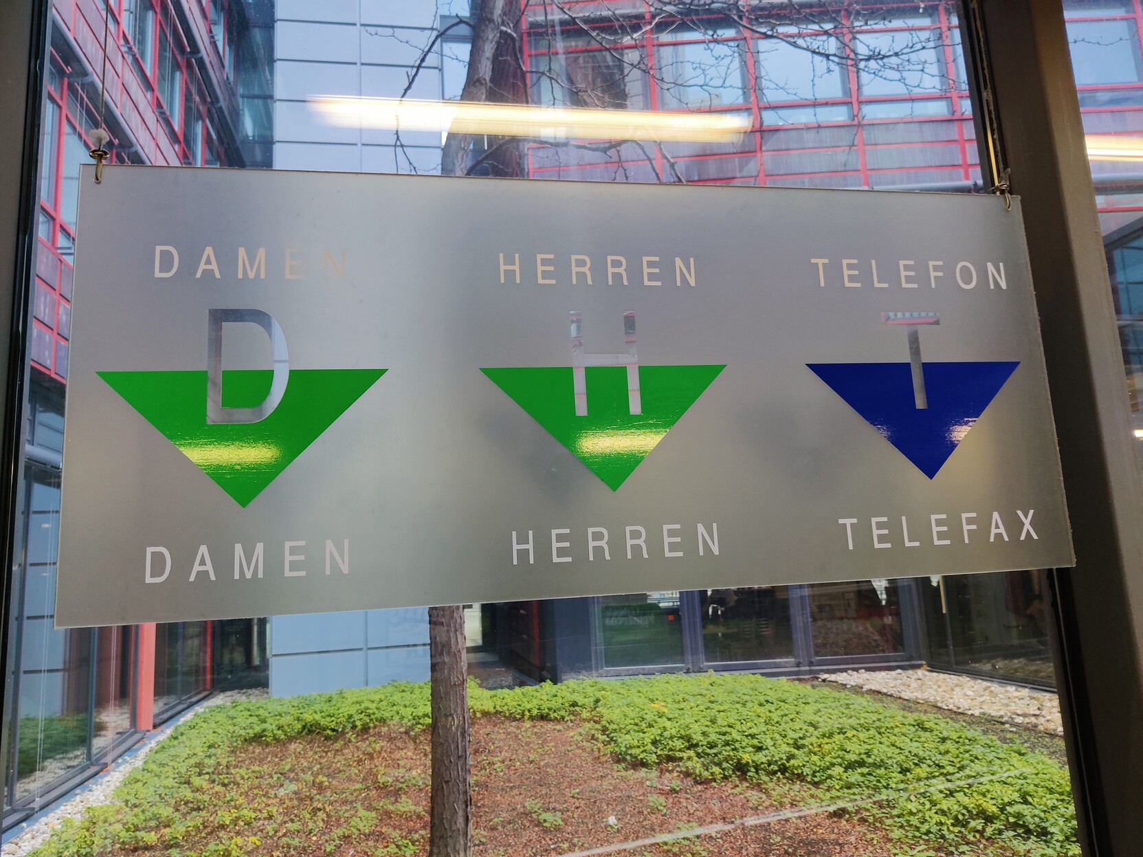 A sign in a public building with three arrows pointing down. The arrows are labeled: DAMEN, HERREN and TELEFON / TELEFAX respectively 