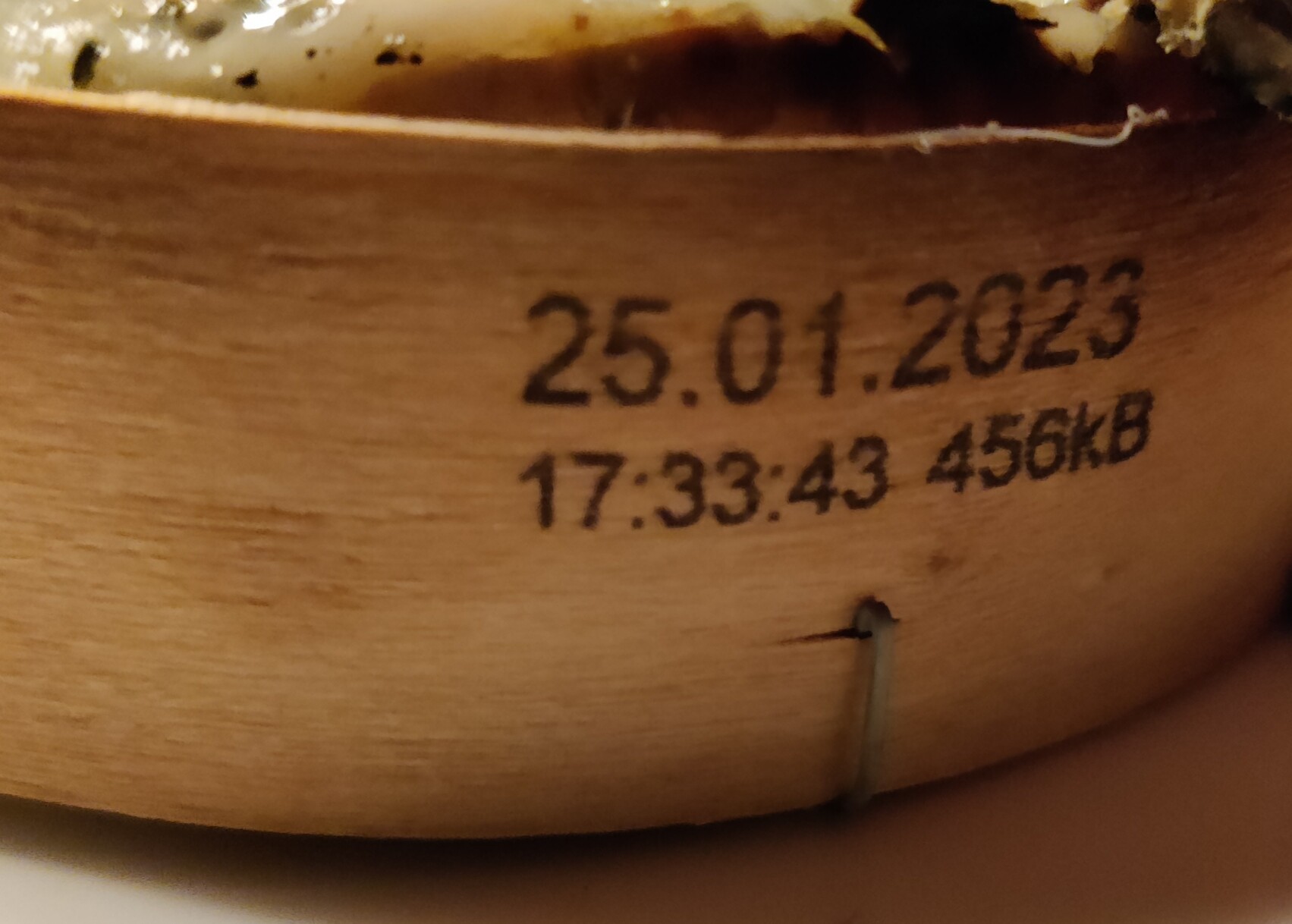 Ein Ofenkäse in der Seitenansicht. Der Holzrahmen ist sichtbar und auf der Seite ist das Mindesthaltbarkeitsdatum 25.01.2023 gedruckt. Darunter die Uhrzeit (vermutlich der Produktion): 17:33:43 und dahinter: 