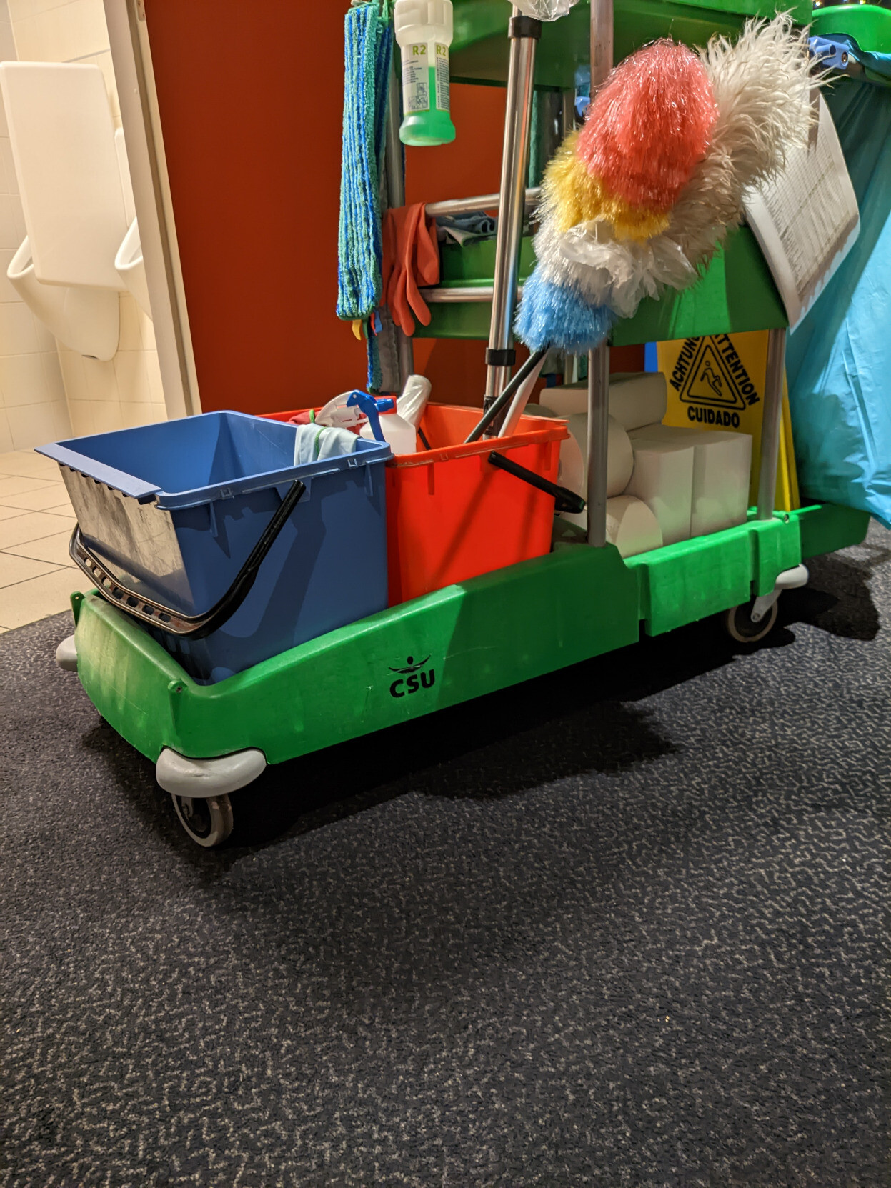 Ein Bild eines Reinigungswagens mit mehreren bunten Eimern und Mops. Auf dem grünen Wagen steht in blauer Schrift 