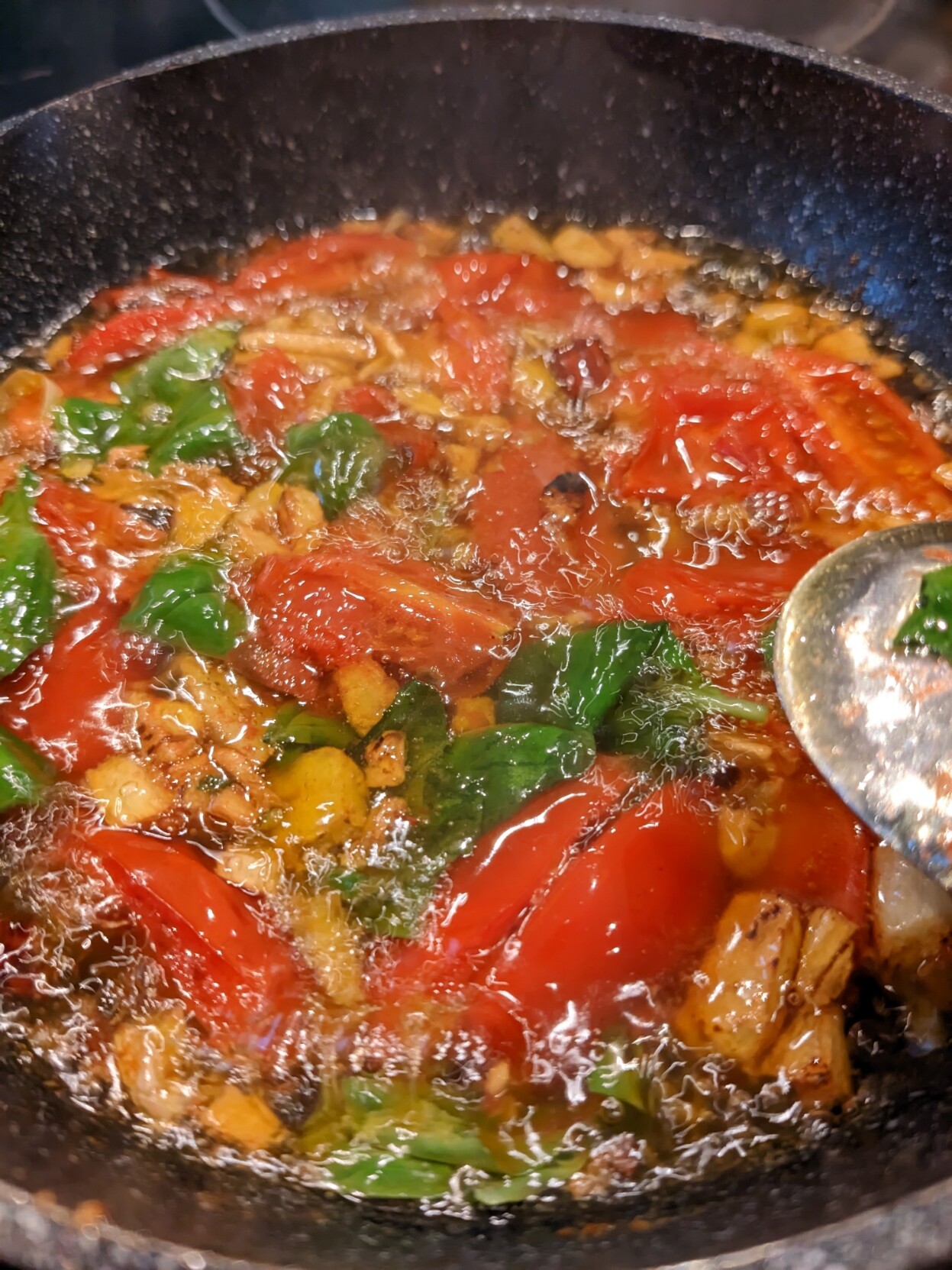 A closeup picture of a small dark frying pan with garlic, tomatoes, basil, sizzling in lots of olive oil.