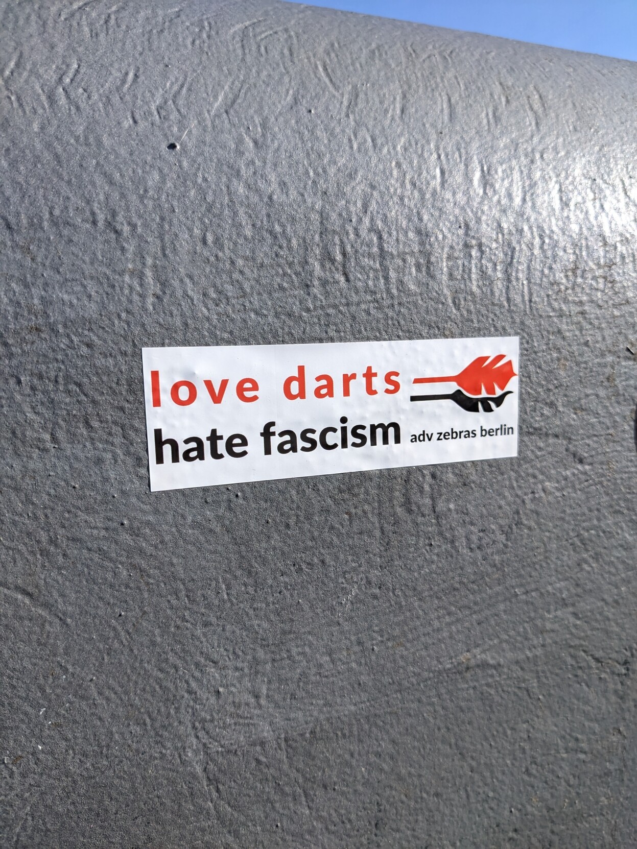 A red and black sticker on a dark grey concrete wall. The sticker reads: 