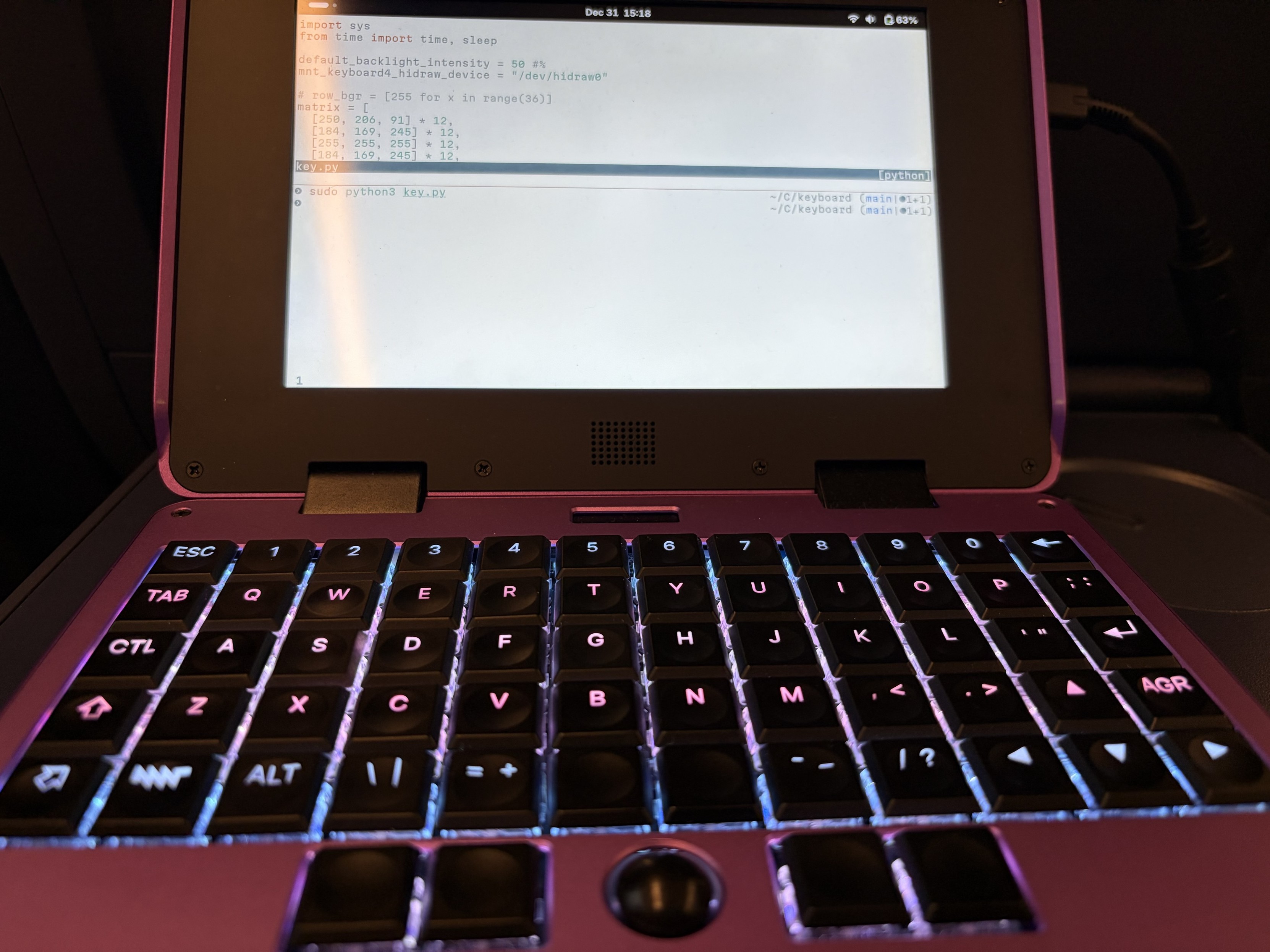 My MNT Pocket Reform on the DB train table. The screen shows a terminal with some Python code, and the keyboard lights up as a transgender flag